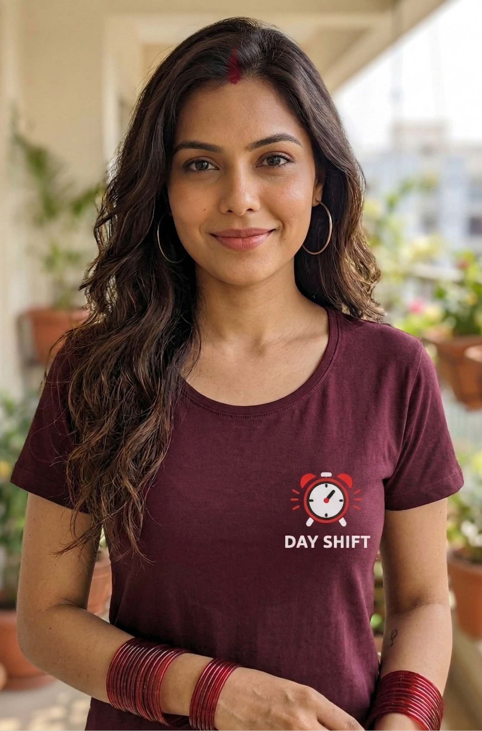 Woman wearing a maroon t-shirt with 'Day Shift' logo on a balcony with plants.