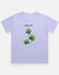 A light lavender kids' t-shirt featuring a green shamrock graphic and the text 'GOOD LUCK' printed on the chest, displayed against a clean off-white background.