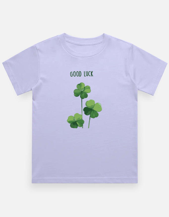 A light lavender kids' t-shirt featuring a green shamrock graphic and the text 'GOOD LUCK' printed on the chest, displayed against a clean off-white background.