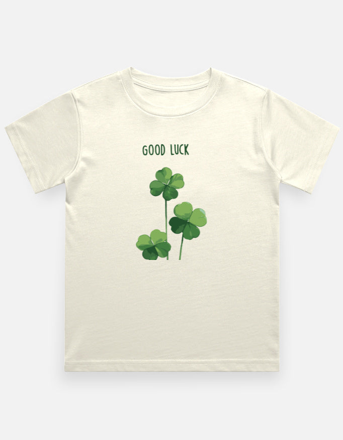 An off-white short-sleeve crewneck t-shirt featuring a green graphic of three shamrocks and the text 'GOOD LUCK' printed on the chest, displayed against a light gray background.