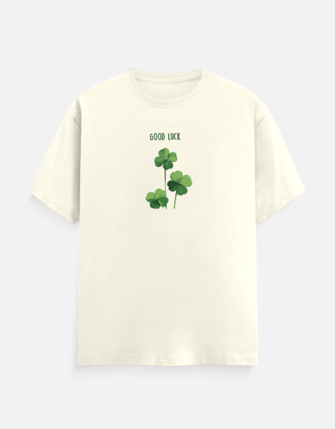 An off-white short-sleeve crewneck t-shirt featuring a green graphic of three shamrocks with the text 'GOOD LUCK' printed above them, displayed on a light gray background.