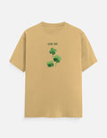 A beige short-sleeve crewneck t-shirt featuring a green shamrock graphic and the text 'GOOD LUCK' printed on the chest, displayed against a light gray background.