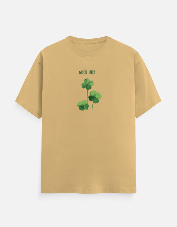 A beige short-sleeve crewneck t-shirt featuring a green shamrock graphic and the text 'GOOD LUCK' printed on the chest, displayed against a light gray background.