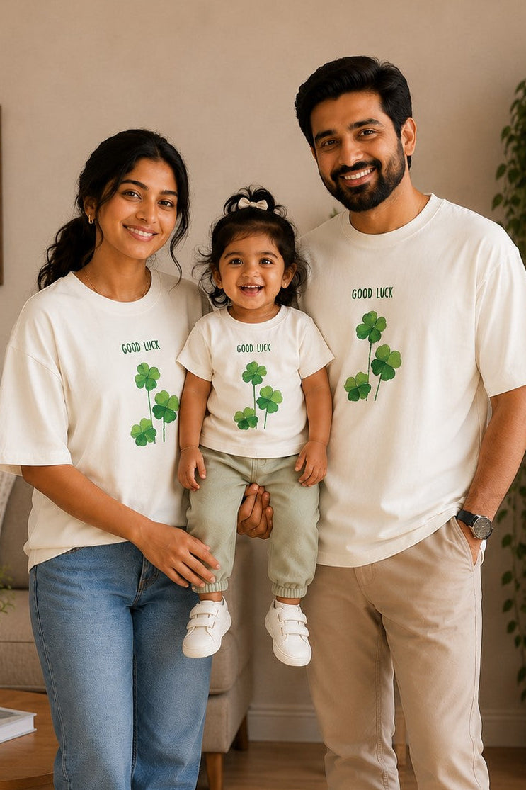 A family of three wearing matching white t-shirts with green shamrock graphics and 'GOOD LUCK' text, standing together in a cozy living room setting.
