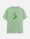 A light green short-sleeve crewneck t-shirt featuring a graphic of three green shamrocks with the text 'GOOD LUCK' printed above them, displayed on a clean white background.