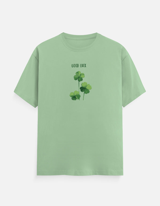 A light green short-sleeve crewneck t-shirt featuring a graphic of three green shamrocks with the text 'GOOD LUCK' printed above them, displayed on a clean white background.
