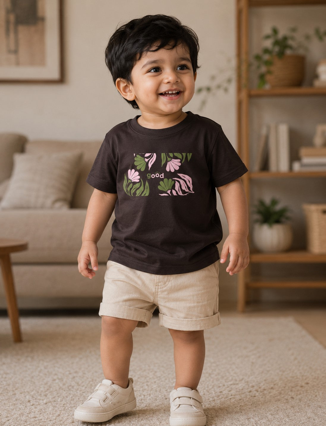 A toddler standing in a cozy living room, wearing a dark brown graphic t-shirt with a pink flamingo and green leaves, paired with beige shorts and white sneakers.