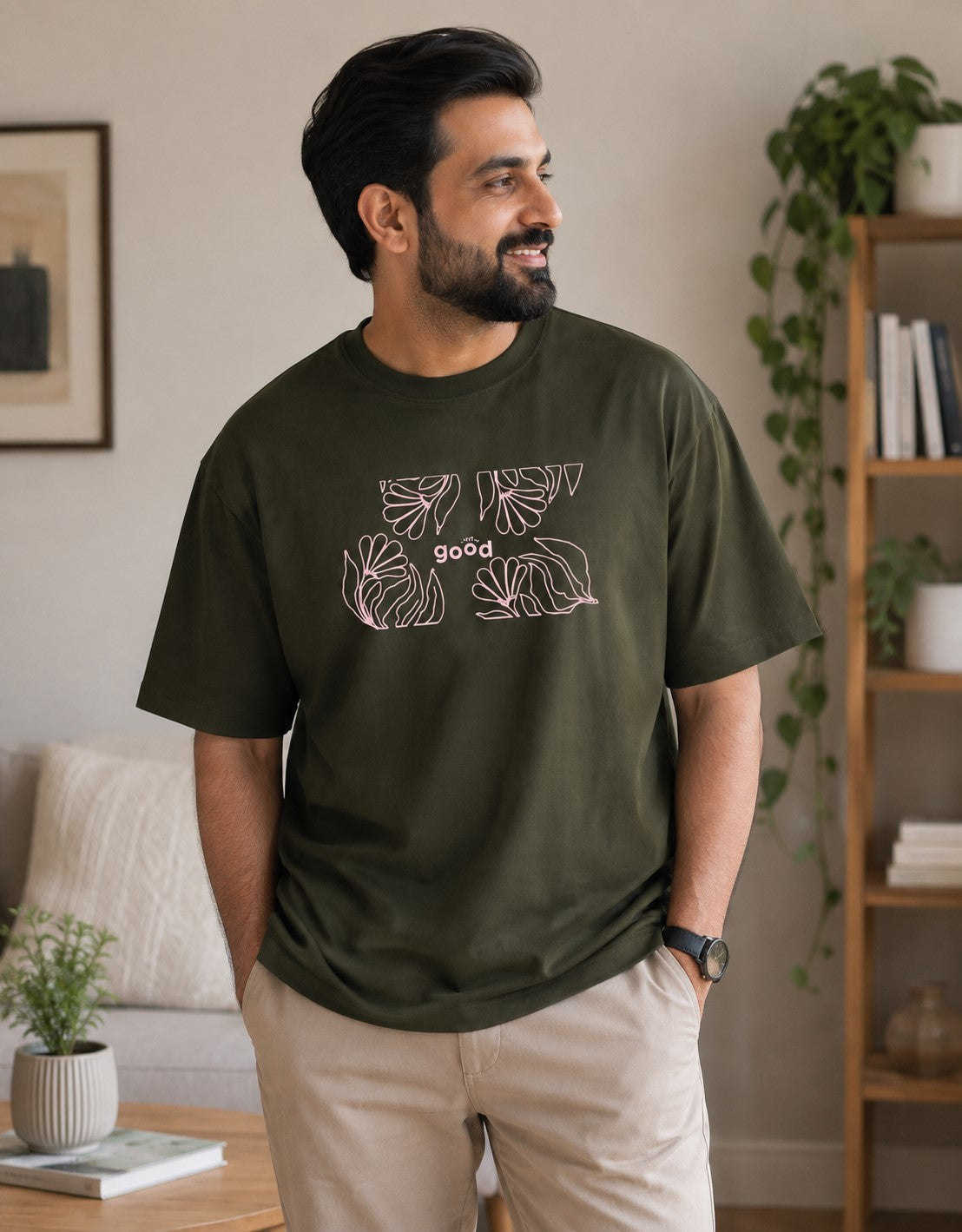 A man wearing an olive green t-shirt with a pink graphic print of monstera leaves and the word 'good' on the front, paired with beige pants, standing in a cozy, modern living room setting.