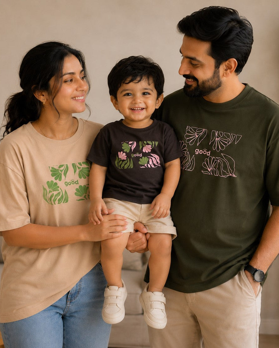 A family of three posing together in a cozy living room setting, each wearing a graphic t-shirt. The woman wears a beige t-shirt with a green leaf print, the young boy wears a dark brown t-shirt with a pink flamingo print, and the man wears an olive green t-shirt with a white leaf graphic.