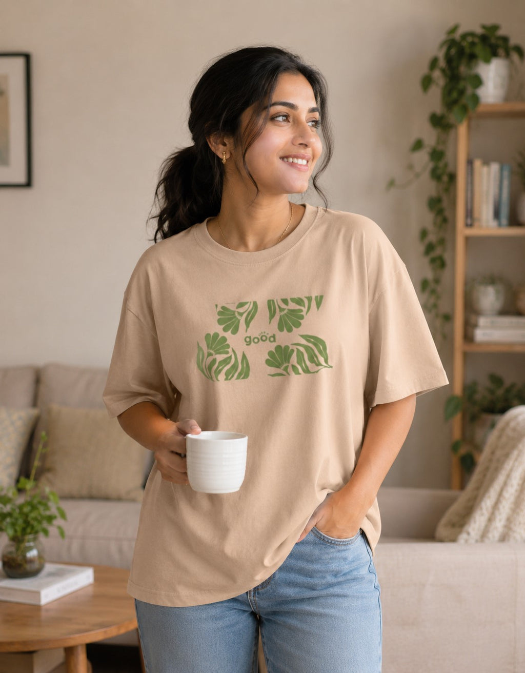 A woman wearing a beige crewneck t-shirt with a green tropical leaf graphic and the word 'goa' printed on the front, holding a white mug in a cozy living room setting.