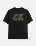 A black short-sleeve crewneck t-shirt featuring a green graphic print of tropical leaves and the word 'good' on the chest, displayed against a light gray background.