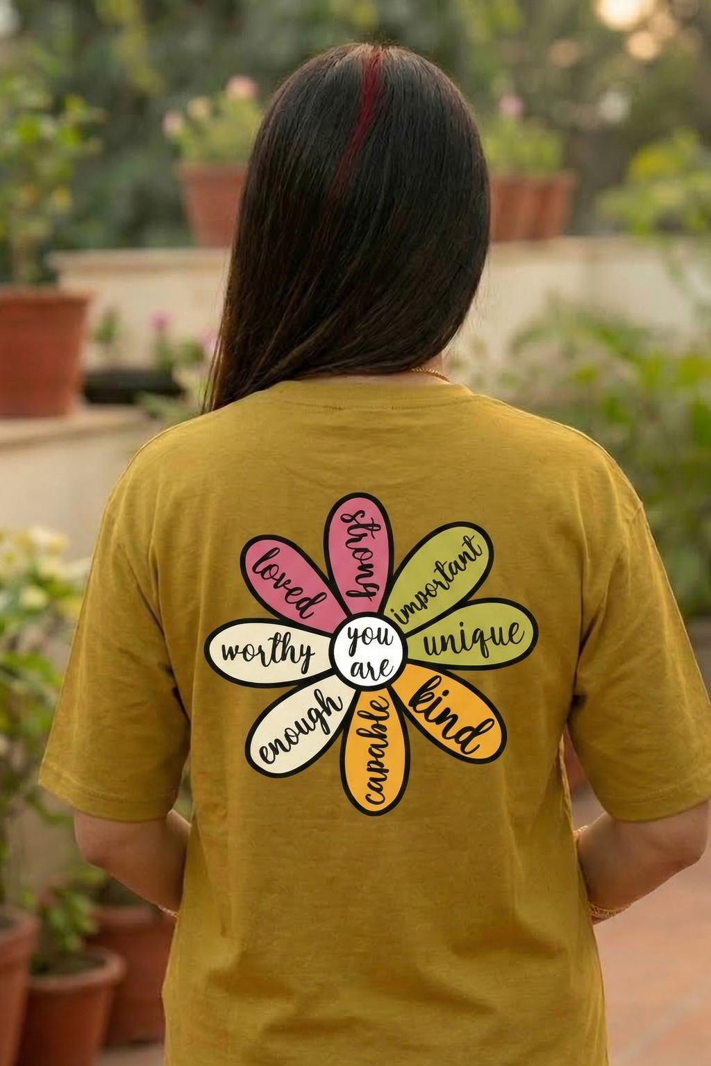 Person wearing a mustard yellow t-shirt with a colourful flower motivational design and text in an outdoor setting.