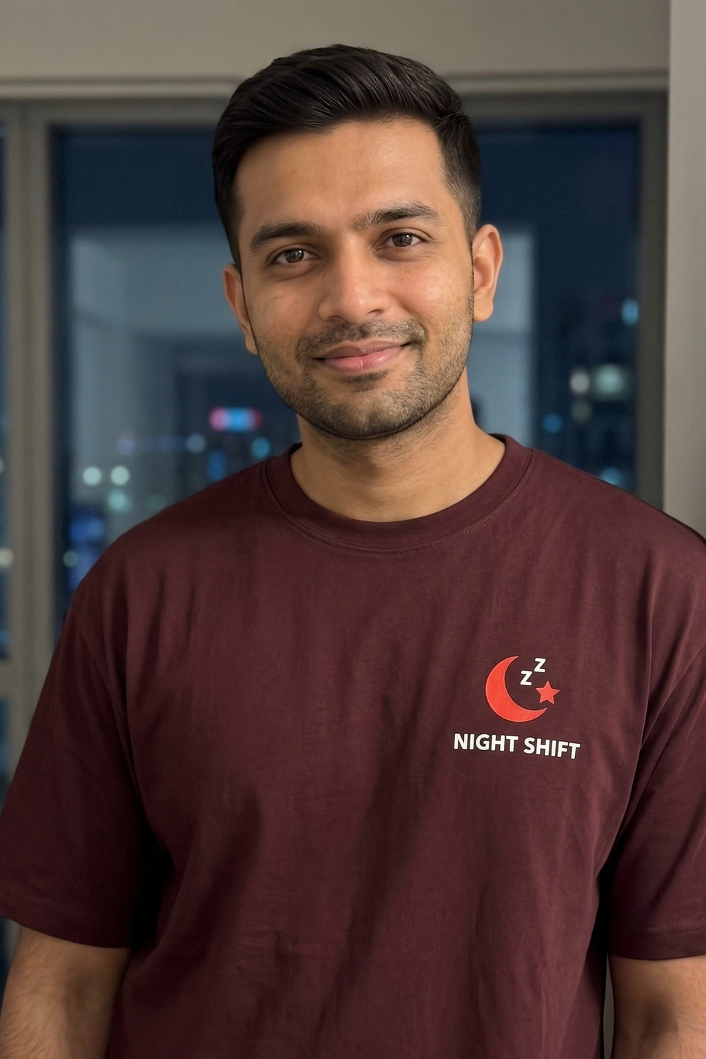 Man wearing a maroon t-shirt with 'Night Shift' logo in front of a cityscape at night.