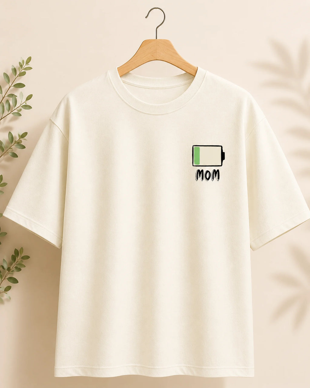 An off-white short-sleeve t-shirt hanging on a wooden hanger against a warm beige background with soft leaf shadows. The shirt features a small graphic on the left chest of a battery icon with a green fill and the word 'MOM' underneath.