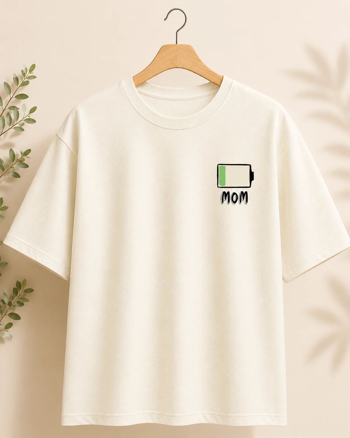 An off-white short-sleeve t-shirt hanging on a wooden hanger against a warm beige background with soft leaf shadows. The shirt features a small graphic on the left chest of a battery icon with a green fill and the word 'MOM' underneath.