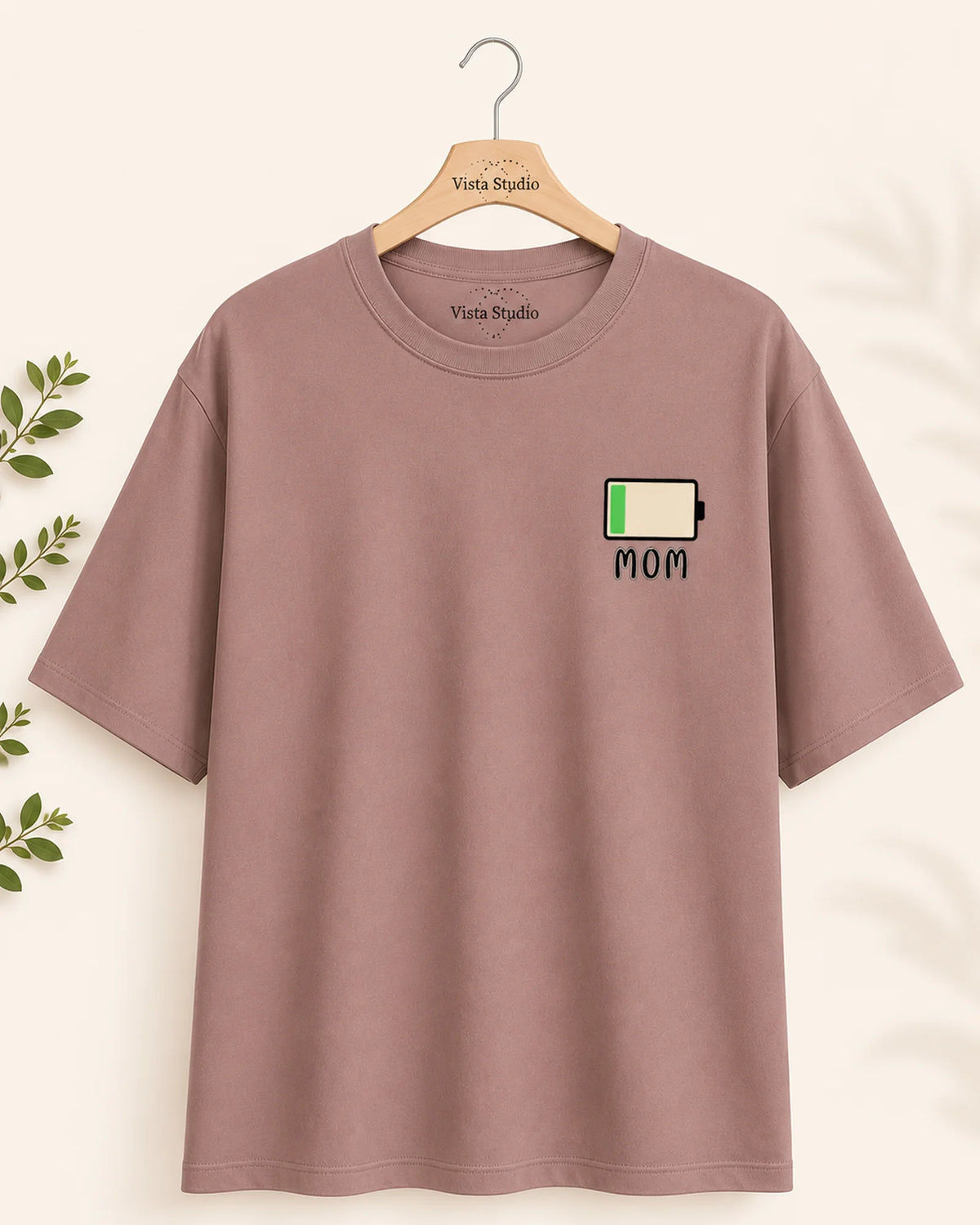 A dusty rose short-sleeve t-shirt hanging on a wooden hanger with 'Vista Studio' printed on it. The shirt features a small graphic on the left chest depicting a battery icon with a green bar and the word 'MOM' underneath.