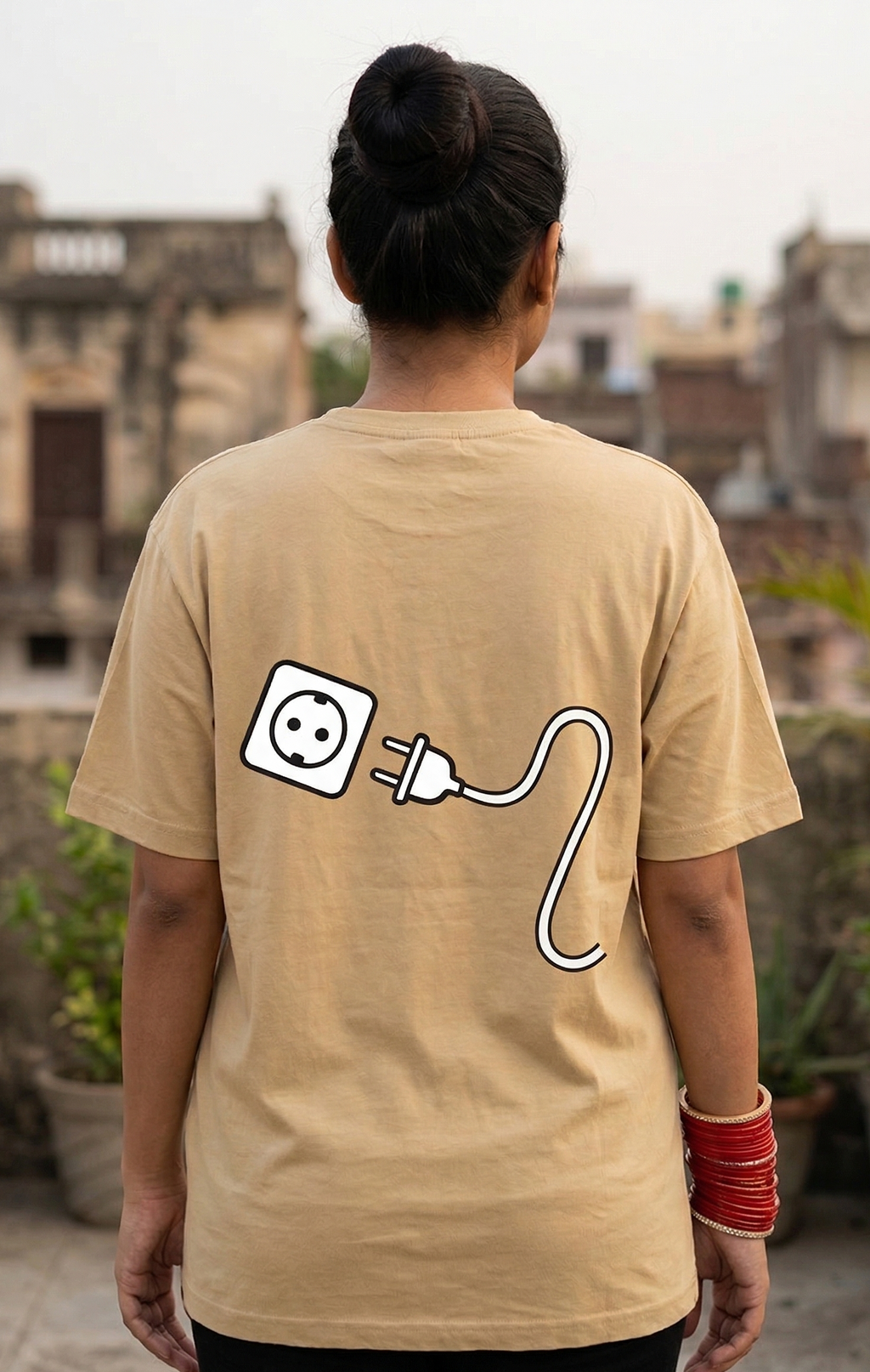 Person wearing a beige t-shirt with a power outlet and cable design on the back, standing on a rooftop with buildings in the background.