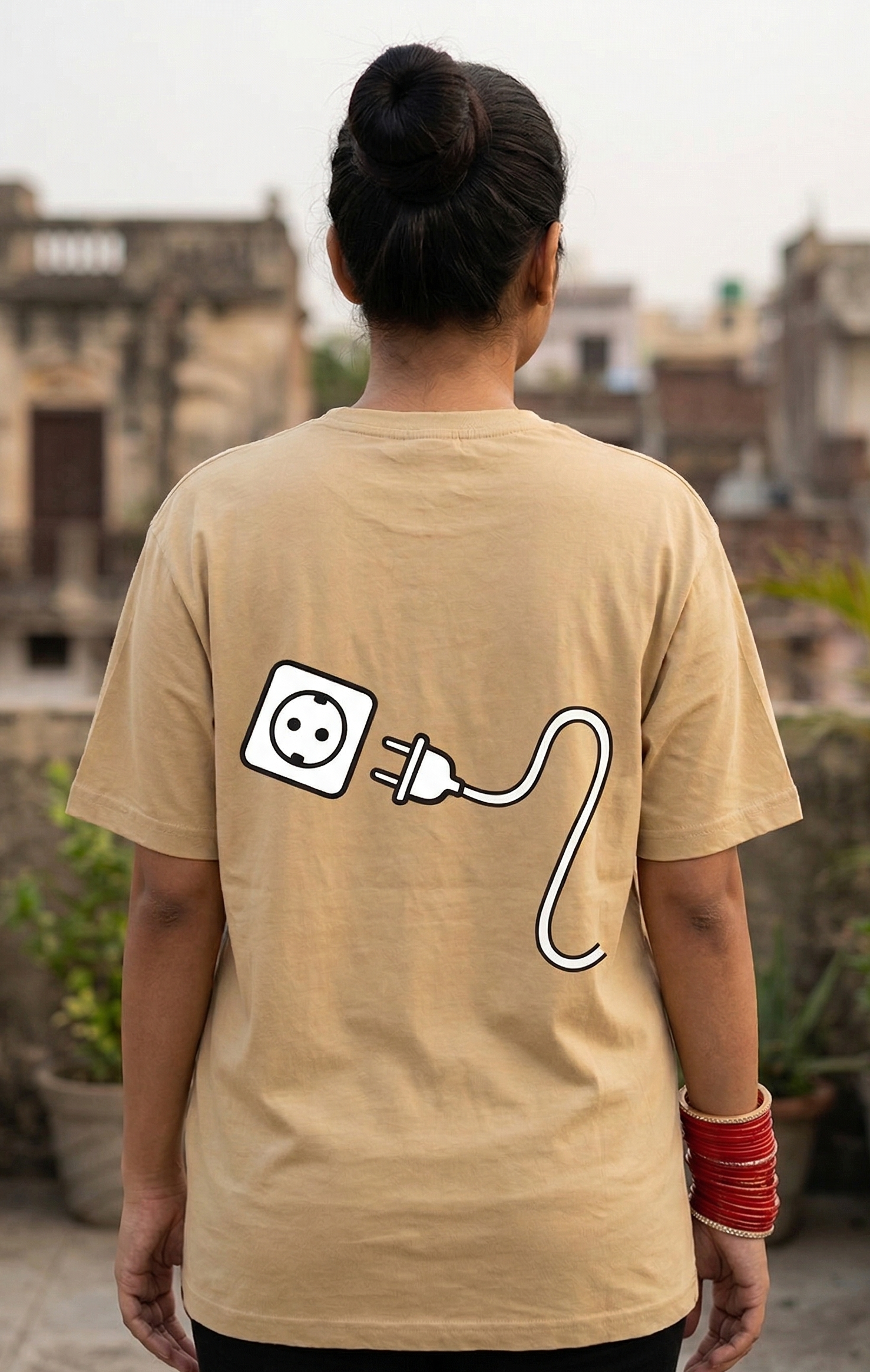 Person wearing a beige t-shirt with a power outlet and cable design on the back, standing on a rooftop with buildings in the background.